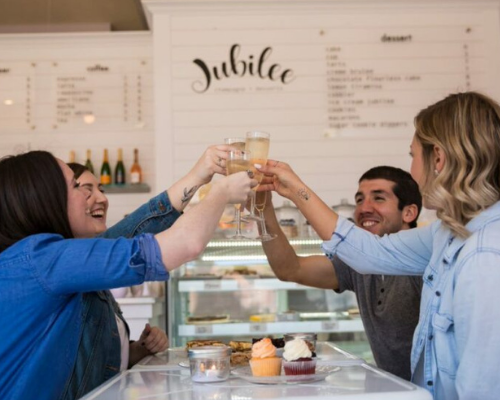 Friends holding up filled champagne glasses cheersing over desserts from from Jubilee